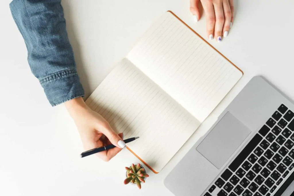 female hand writing notebook table with laptop scaled